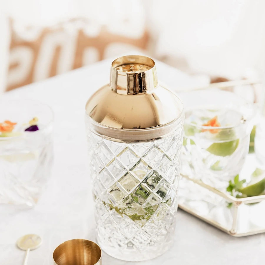 lifestyle photo of cut glass cocktail shaker with gold strainer on white table with lid alongside