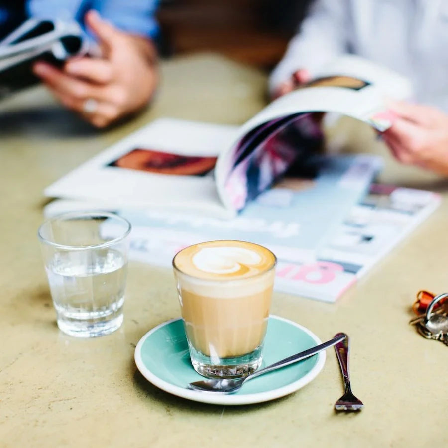lifestyle photo of coffee shop with latte in coffee glass on green plate on table with glass of water and man reading magazine